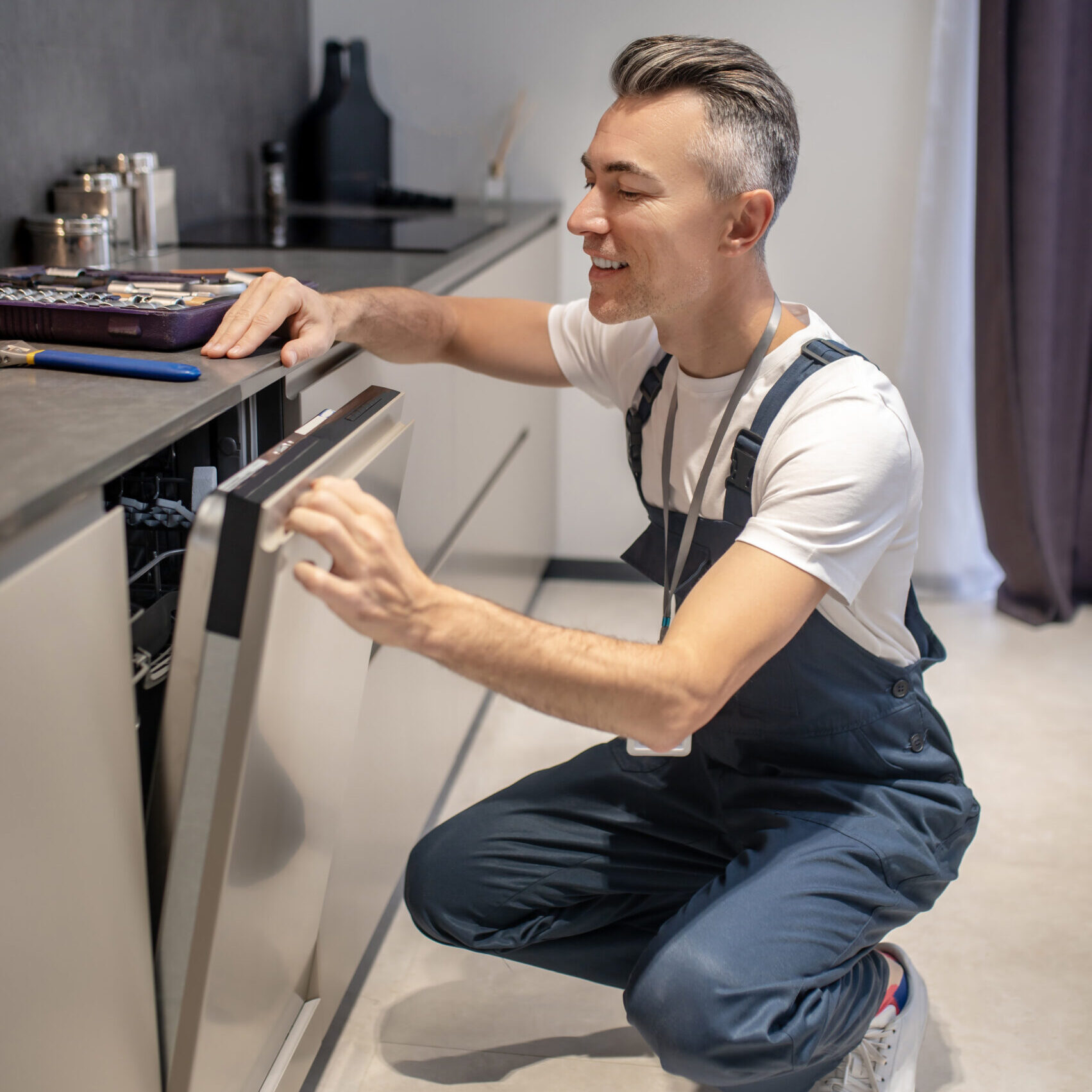 Working mood. Smiling man in tshirt and overalls crouching peeking into open door of built-in dishwasher in modern kitchen